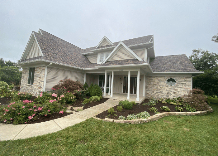 Elegant two-story house with a gabled roof, featuring stone and siding exterior, surrounded by landscaped garden with pink flowers.