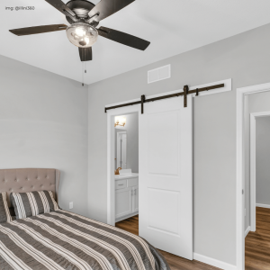 A stylish bedroom with gray walls and a sliding white barn door leading to the en suite bathroom. The room features a taupe tufted headboard, a striped comforter, and a dark ceiling fan with a light fixture. The floor is hardwood, and there is a view into the white-trimmed bathroom with a golden faucet.