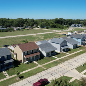 An aerial view of a suburban neighborhood featuring a variety of single-family homes with diverse roofing and siding colors. The neatly organized homes have well-maintained lawns and are set along a quiet street, with spacious gaps and green areas around them.