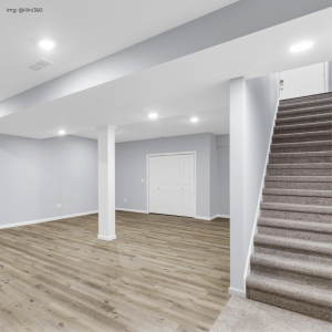 A spacious and modern basement area featuring soft gray walls, recessed lighting, and elegant wood flooring. The space is highlighted by a white column and includes a carpeted staircase leading upstairs, with a white door that presumably leads to additional rooms.