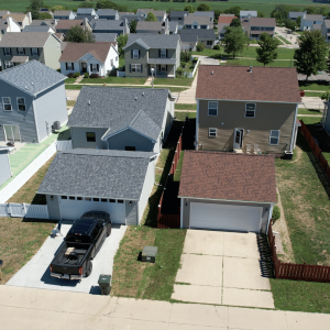 An aerial view of a suburban neighborhood showing a mix of single and two-story homes with various siding colors. The homes have well-kept lawns, driveways with vehicles, and fenced backyards, set in a neatly planned residential area.