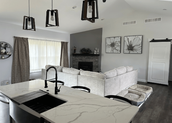 Modern living room with an open floor plan featuring a large light gray sectional sofa, a stone fireplace with decor, and a sleek black kitchen island faucet in the foreground. The space is well-lit with large windows, hanging pendant lights, and neutral gray tones throughout. Two floral art pieces and a sliding barn door enhance the room's contemporary design.