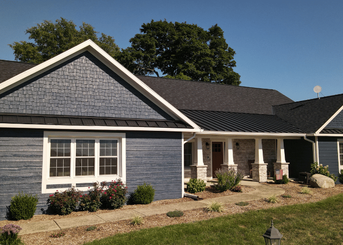 The image shows a modern single-story home with blue siding, white trim, and a dark asphalt shingle roof. The front yard is landscaped with shrubs and flowers, adding to the home’s curb appeal. Ideal for showcasing residential roofing or construction work.