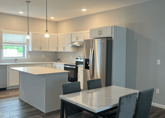 Modern kitchen with white cabinets, stainless steel appliances, and a central island. Features recessed lighting, pendant lights, and a dining area with a marble-style table and upholstered chairs.