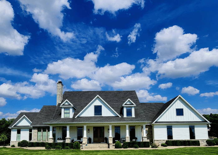 Modern farmhouse-style home with white board and batten siding, black-framed windows, and a dark gray shingle roof under a bright blue sky with scattered clouds.