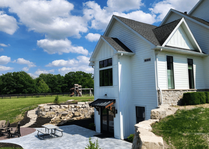 Modern white farmhouse with stone landscaping, outdoor patio, and well-maintained roofing and siding under a sunny sky.