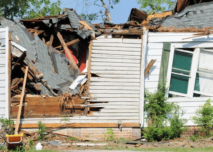 Severe storm damage to a house with a collapsed roof, broken siding, and leaning windows, exposing the interior.