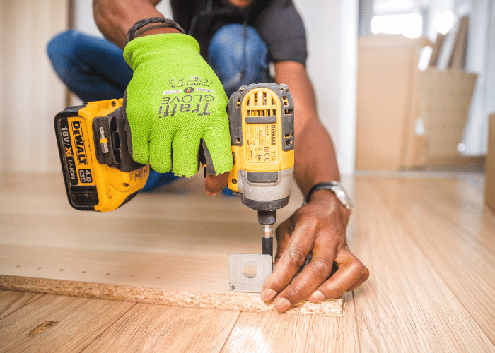 Close-up of a person using a DeWalt power drill to fasten a metal bracket onto wood, wearing a bright green work glove on one hand.